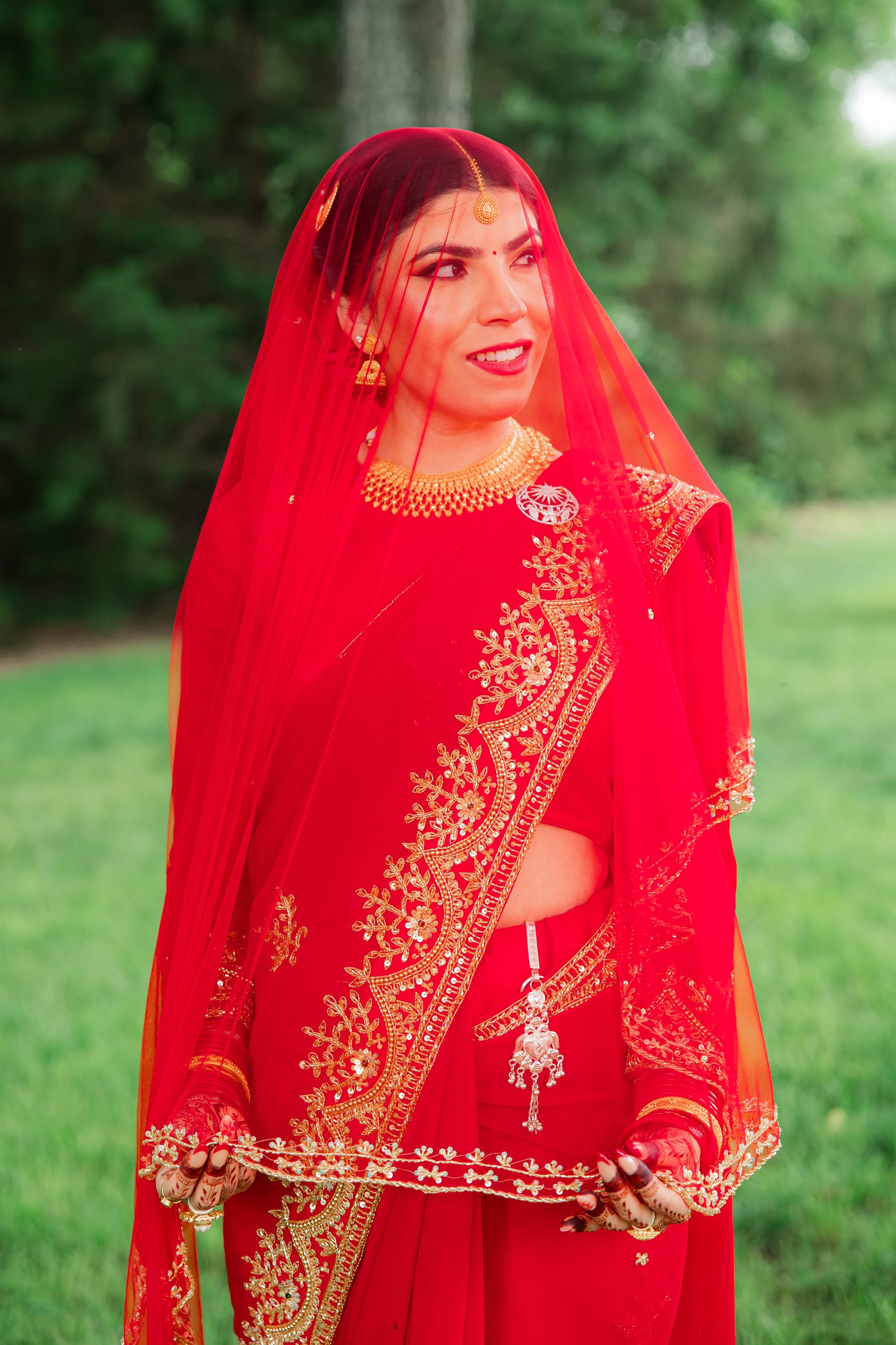 A bride in a red and gold veil and saree smiles while looking off to the side outdoors.