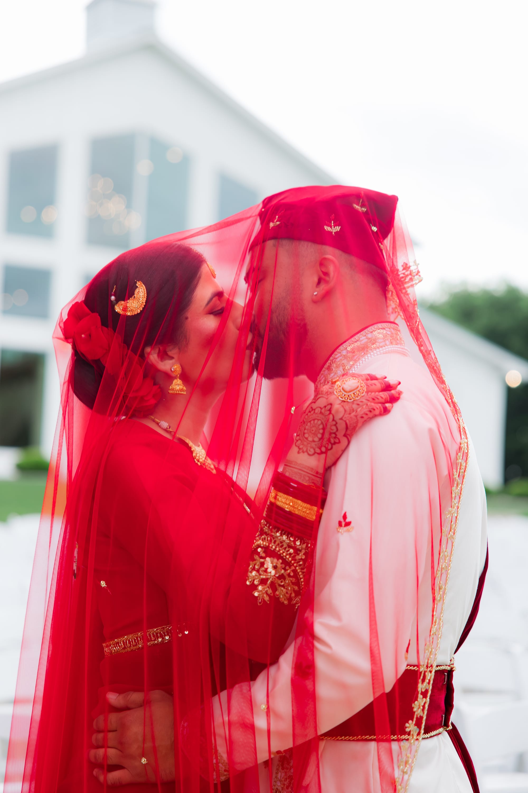 A bride and groom kiss under a sheer red veil held over both their heads outdoors.