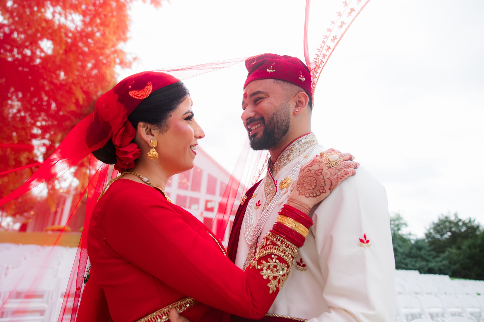 A bride and groom in red and white wedding attire smile at each other under a fluttering red canopy.