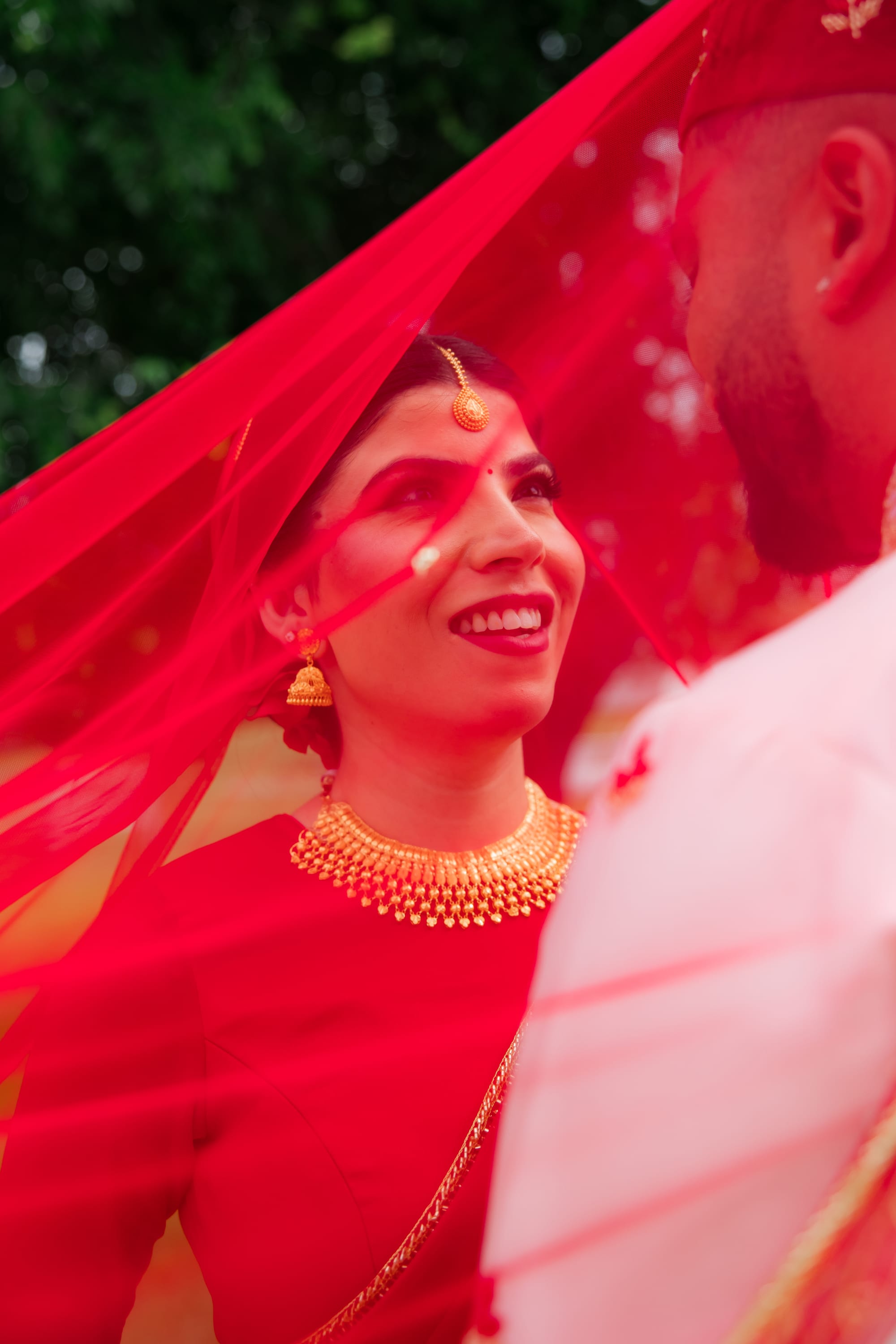 A smiling bride in a red veil and gold jewelry looks up at the groom beside her.