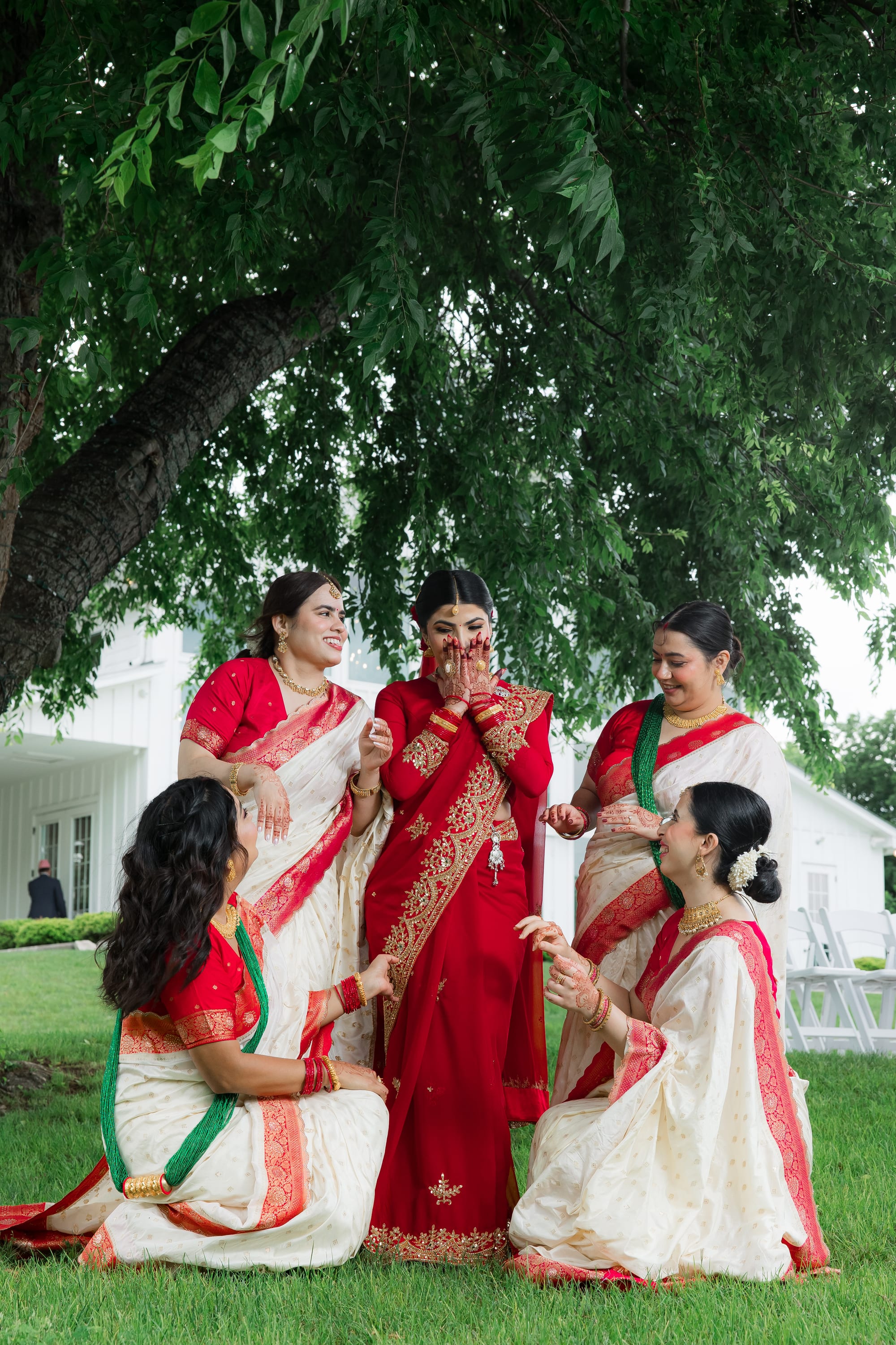 Bridesmaids in cream and red saris surround a laughing bride in a red and gold outfit under a tree.
