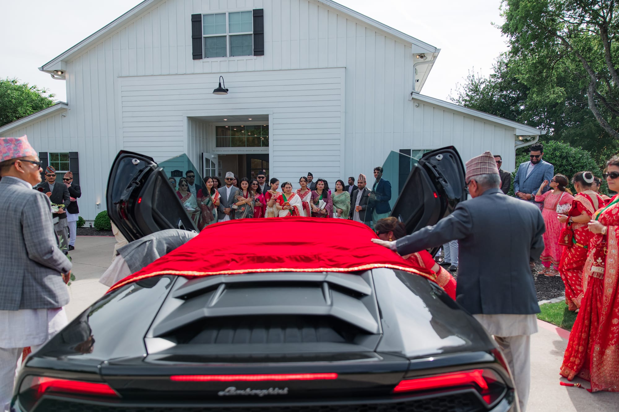 Wedding guests in traditional dress gather around a black sports car with its doors open outside a barn-style venue.