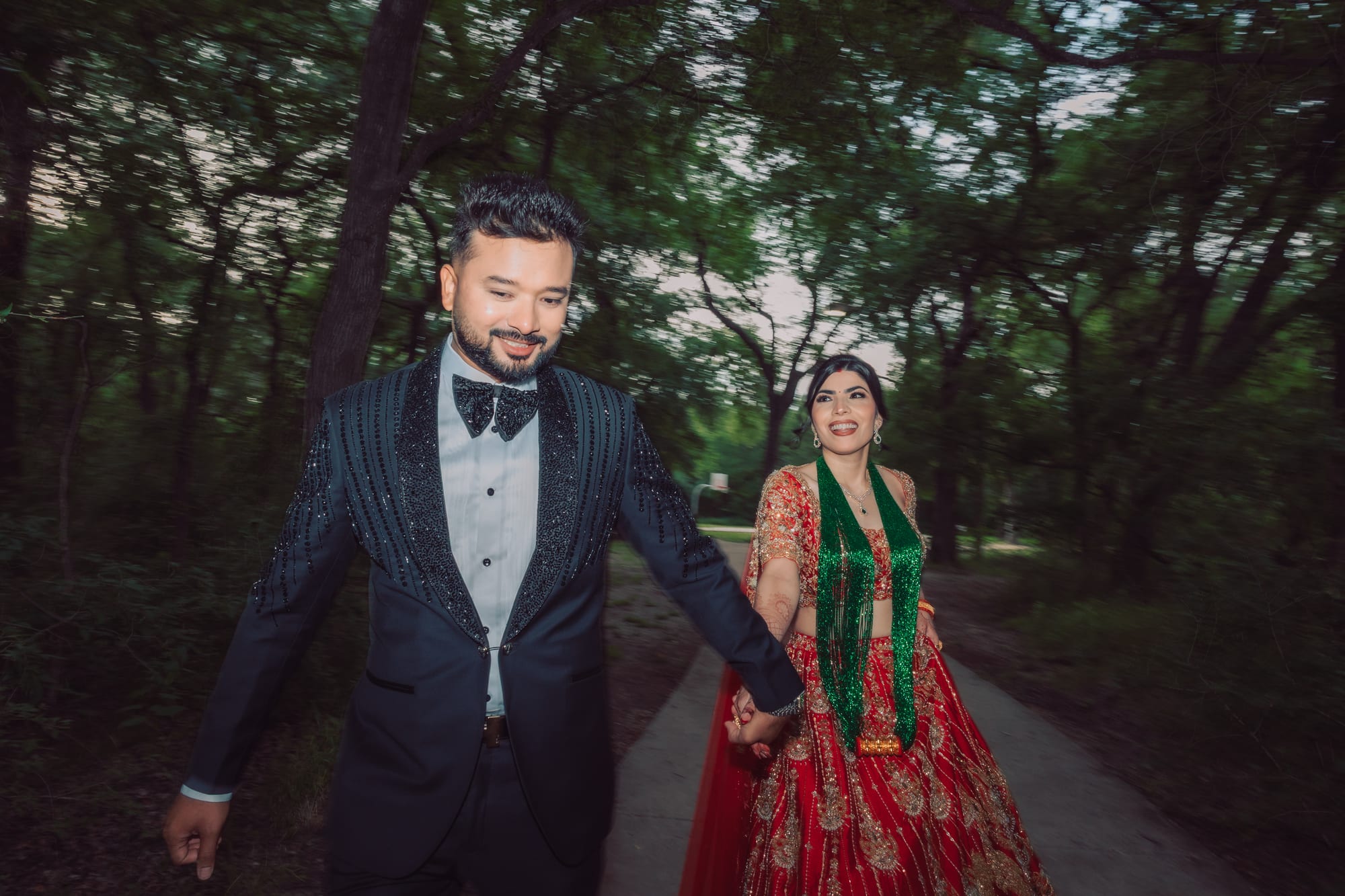 A groom in a beaded tuxedo jacket and a bride in a red and green lehenga walk hand in hand along a wooded path.