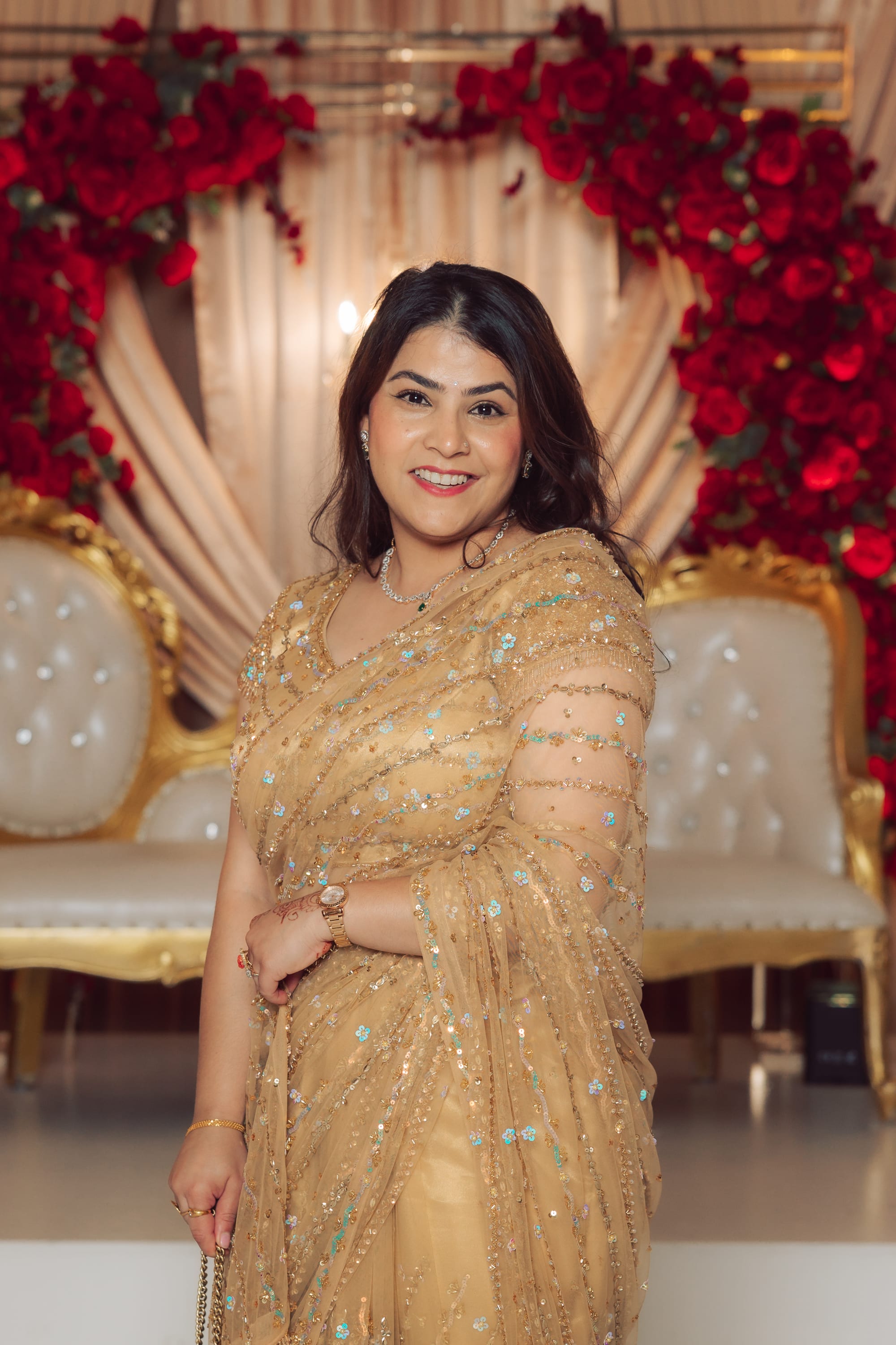A woman in a gold sequined sari smiles in front of a rose-covered arch and gold chairs at an event.
