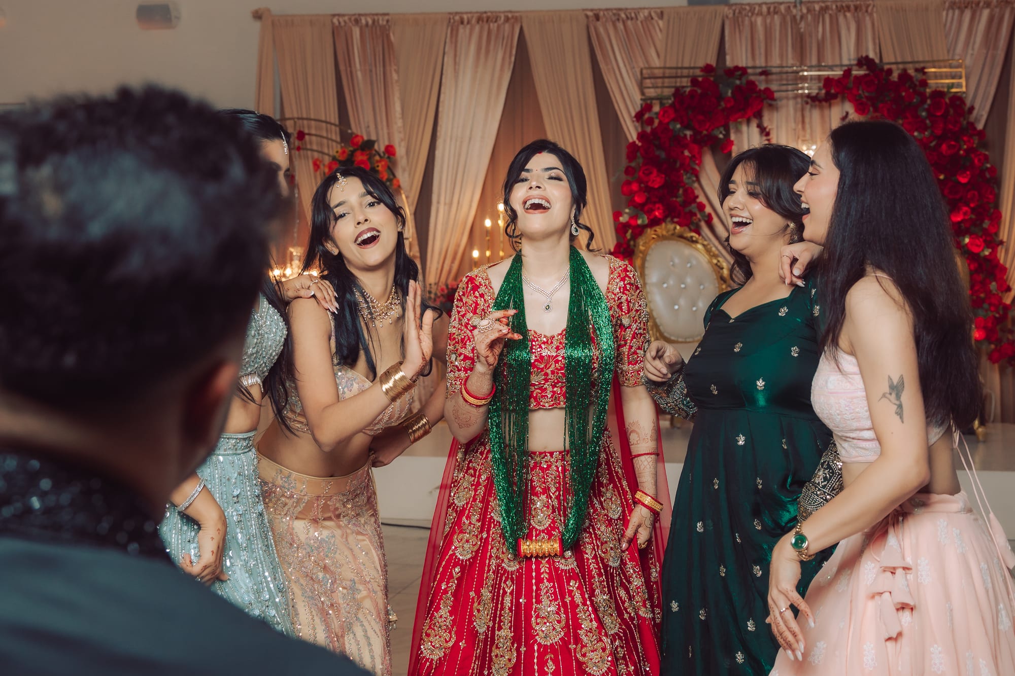 A group of women in colorful dresses laugh together around a bride in a red and green lehenga.