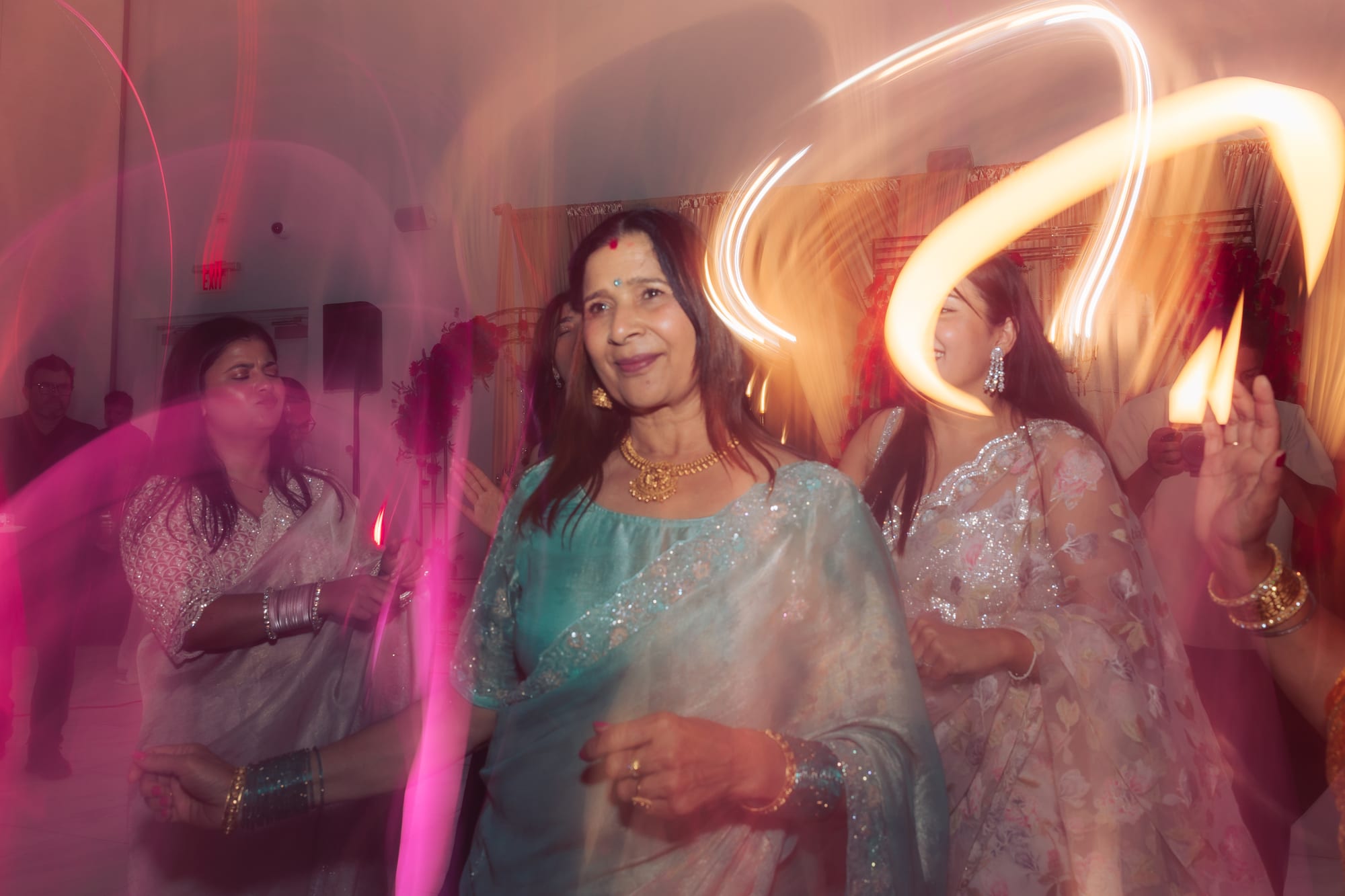 A motion-blurred shot of an older woman in a teal sari dancing amid trailing string lights at a party.