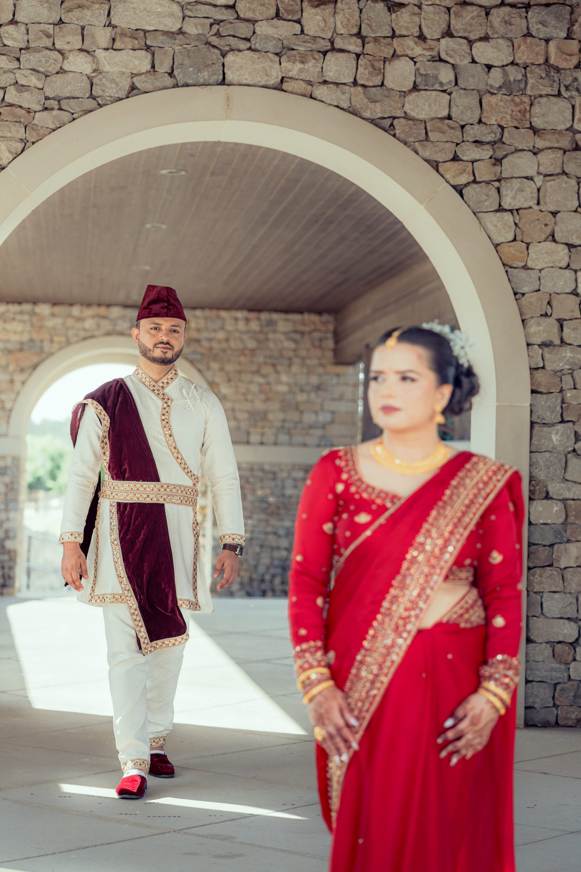 A man in traditional white and maroon dress walks under a stone archway behind a woman in a red saree looking away.
