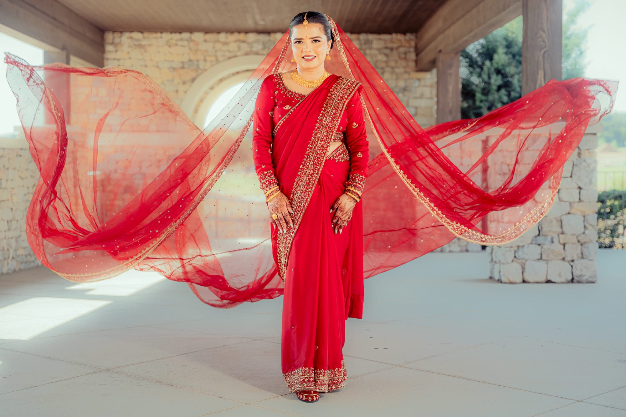 A bride in a red and gold saree stands with her veil caught mid-twirl, spread out on either side of her like wings.