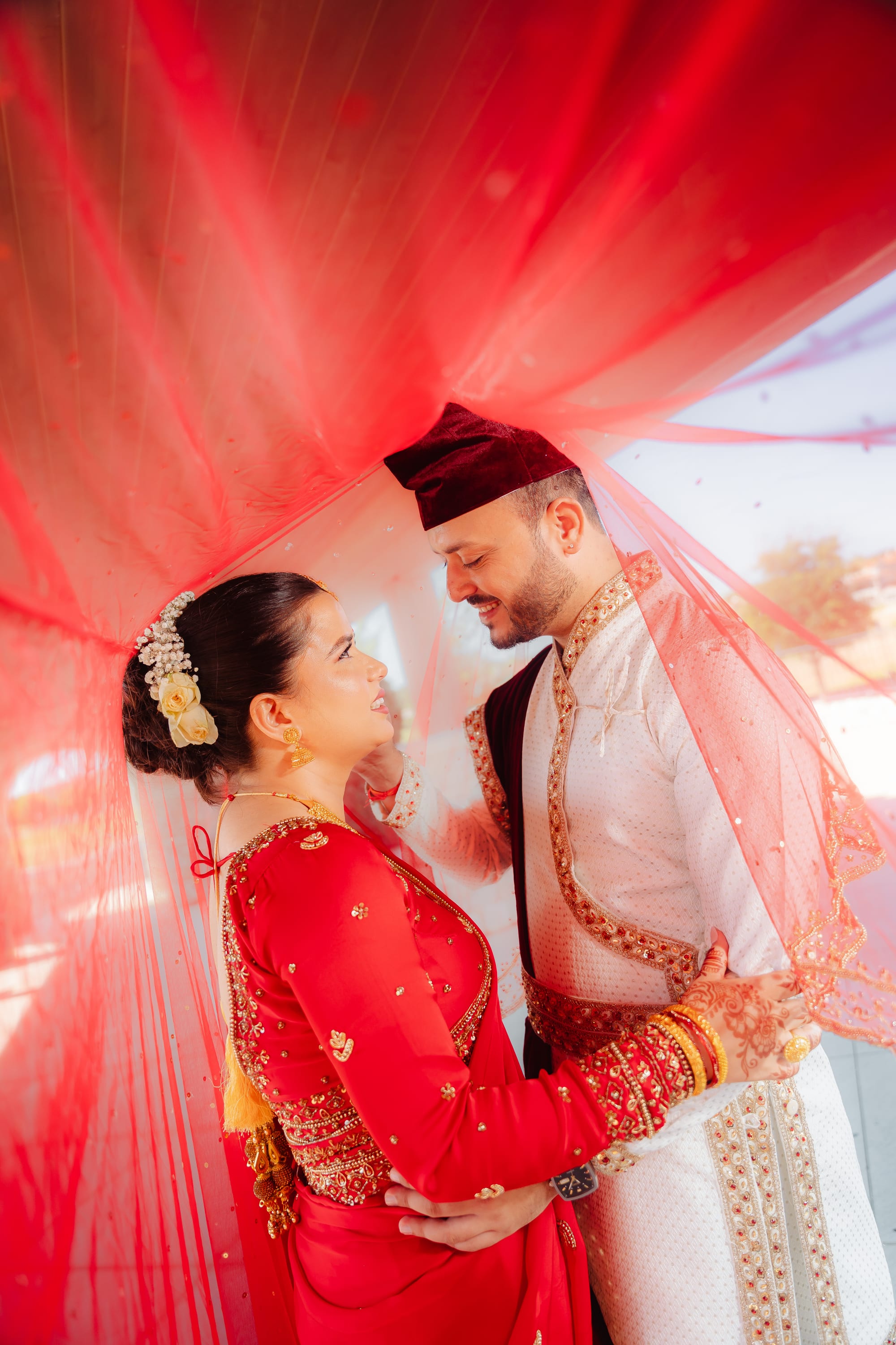 A bride and groom embrace under a red canopy, foreheads close together, both smiling.