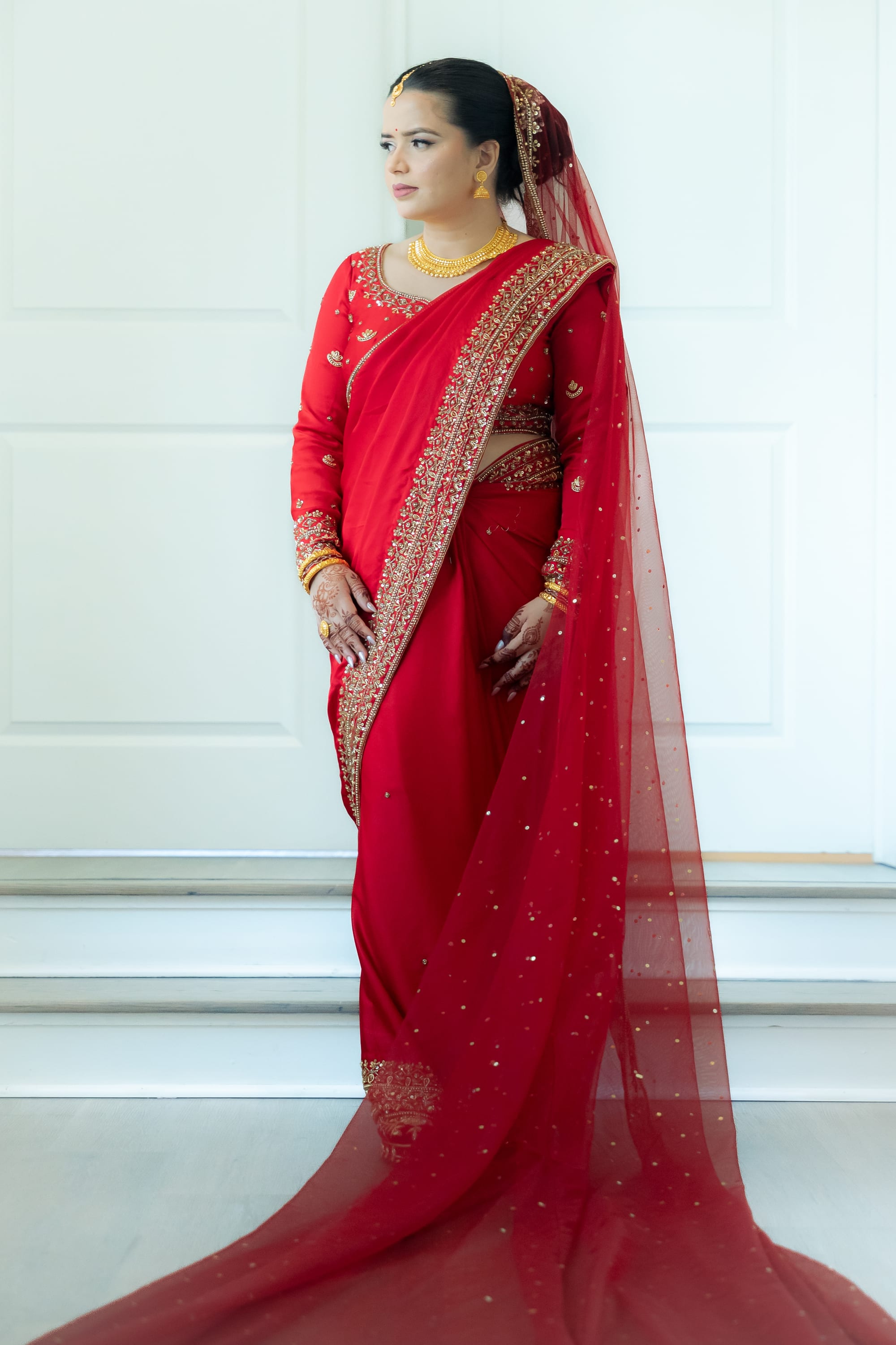 A bride in a red sari with gold embroidery stands indoors, a sheer veil trailing to the floor.
