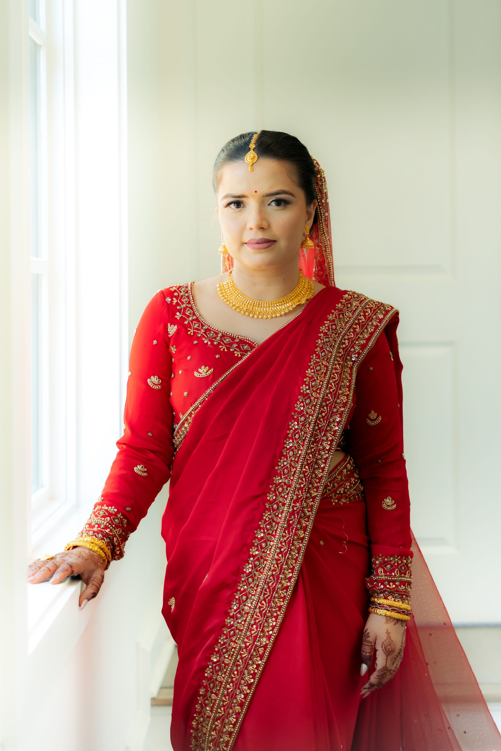 A bride in a red sari and gold jewelry looks at the camera beside a bright window.