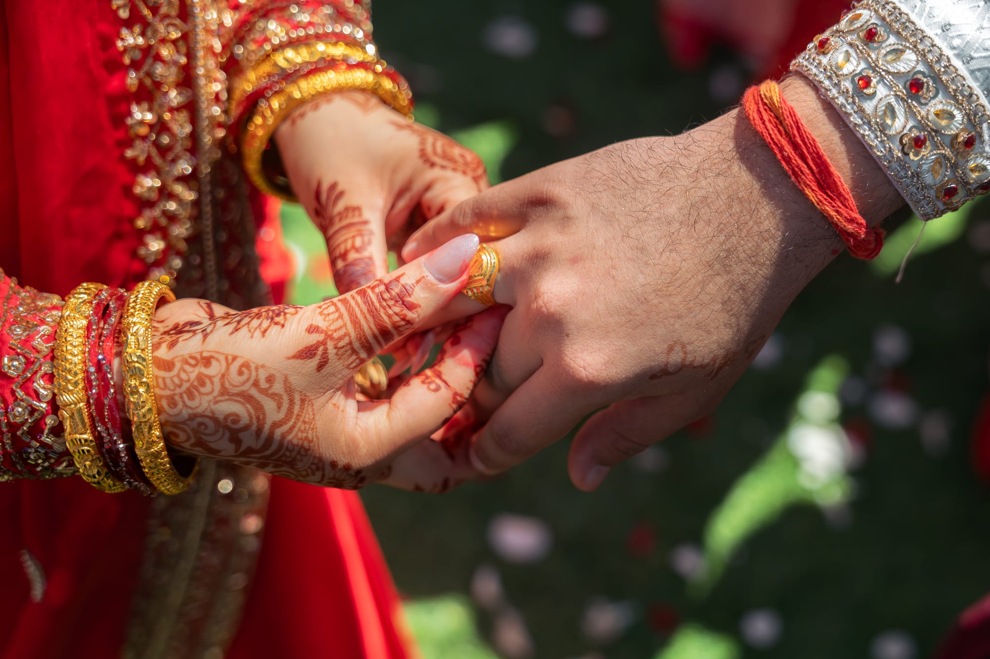 Close-up of a bride and groom's hennaed hands clasped together outdoors, wearing gold rings and bangles.