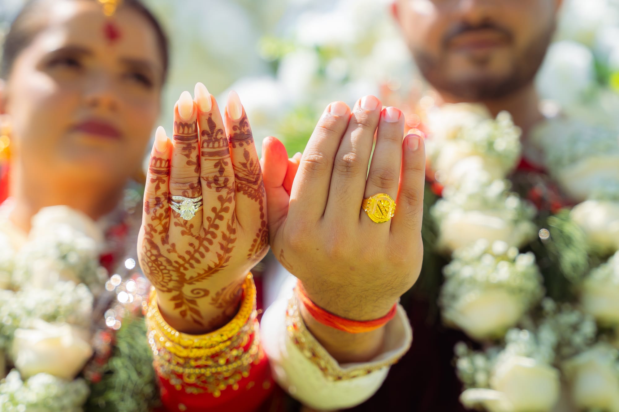 A bride and groom raise their ringed hands side by side in front of white floral garlands.