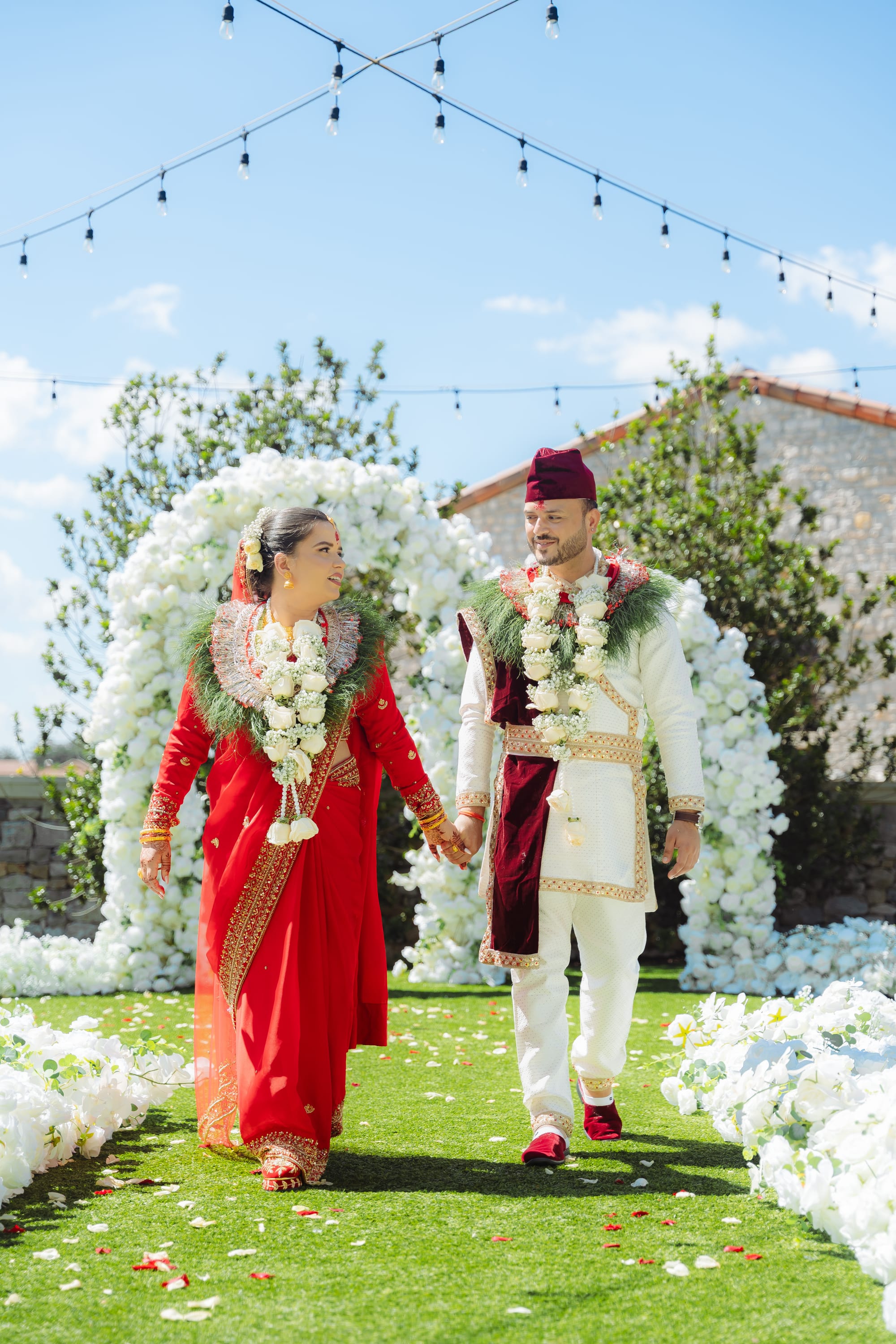 A bride in red and a groom in white and maroon walk hand in hand down a petal-strewn aisle lined with white floral arches.