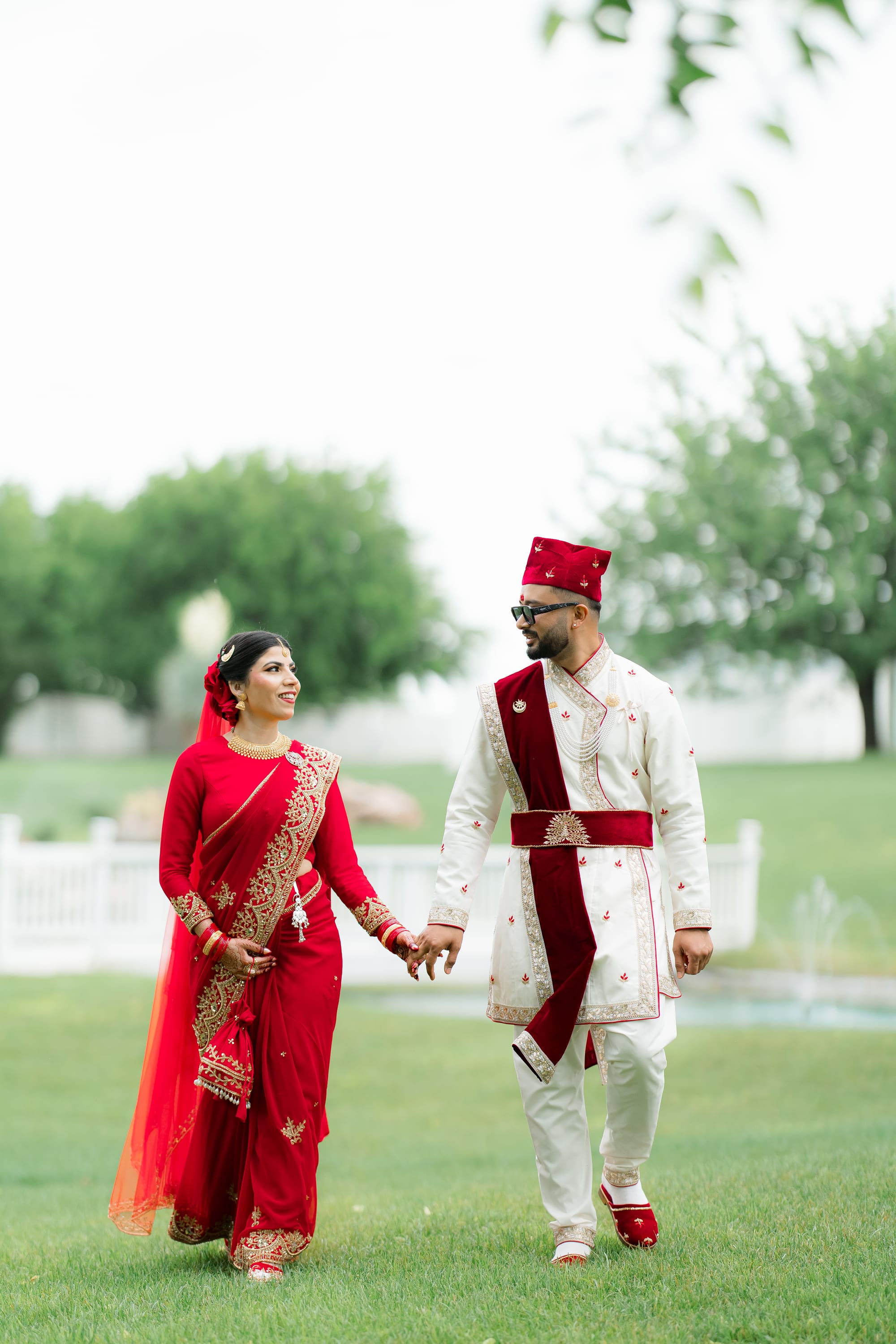 A bride in a red and gold saree and a groom in white and maroon walk hand in hand across a lawn.