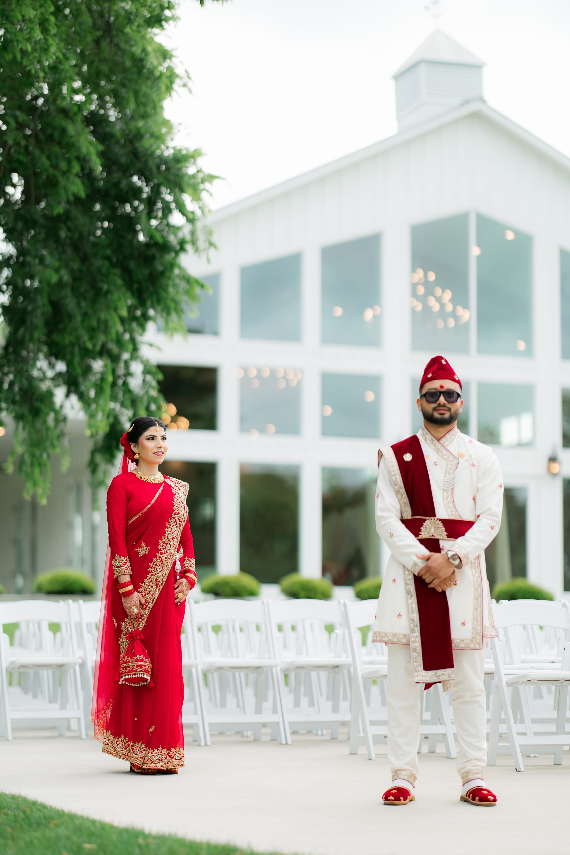 A bride in a red embroidered saree and a groom in white with a red sash smile at each other in a garden.