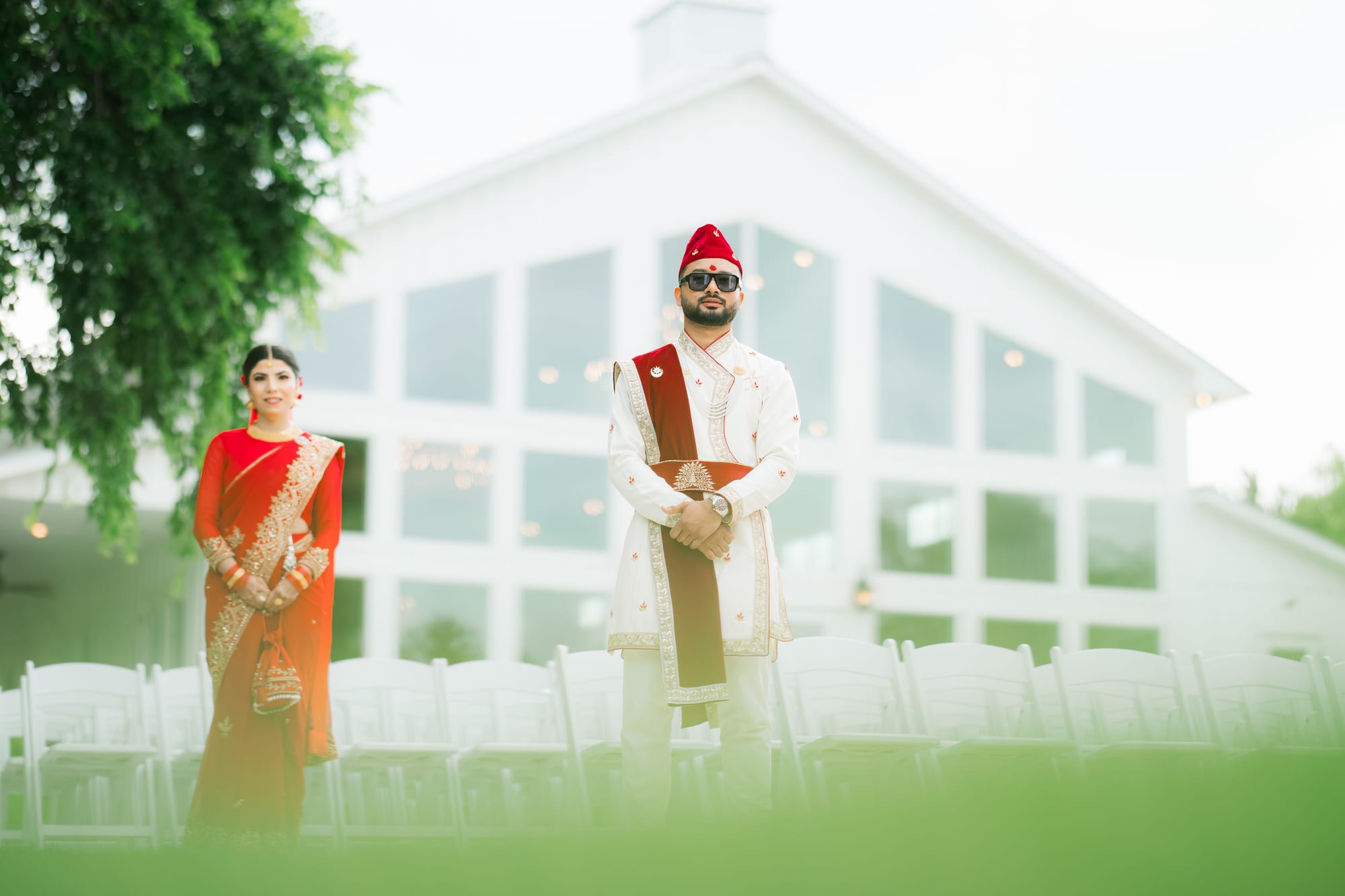 A bride in red and a groom in white and maroon stand apart outside a glass-walled venue at dusk.