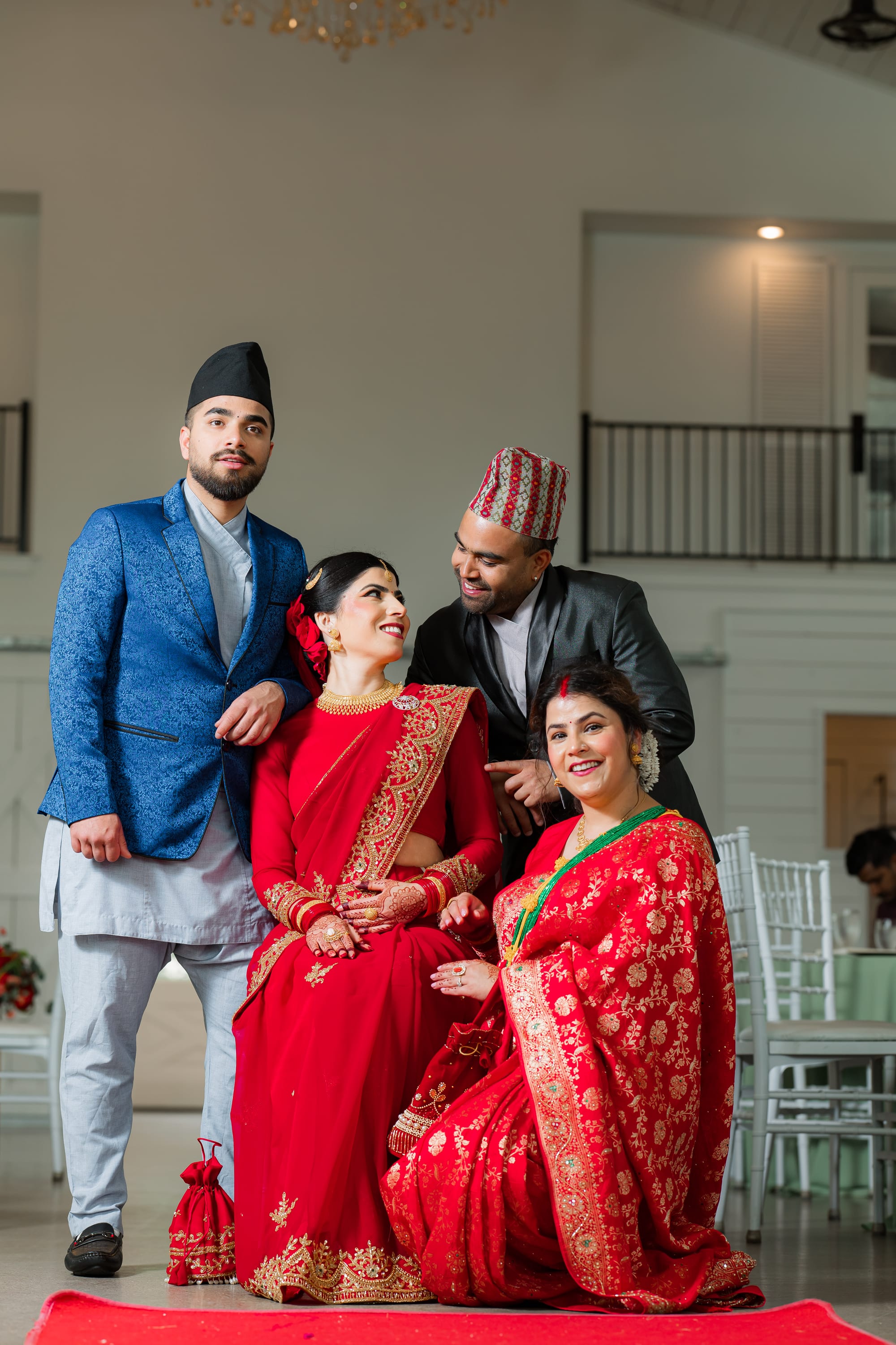 Two men in jackets and topi hats stand behind two seated women in red bridal saris, all smiling together.