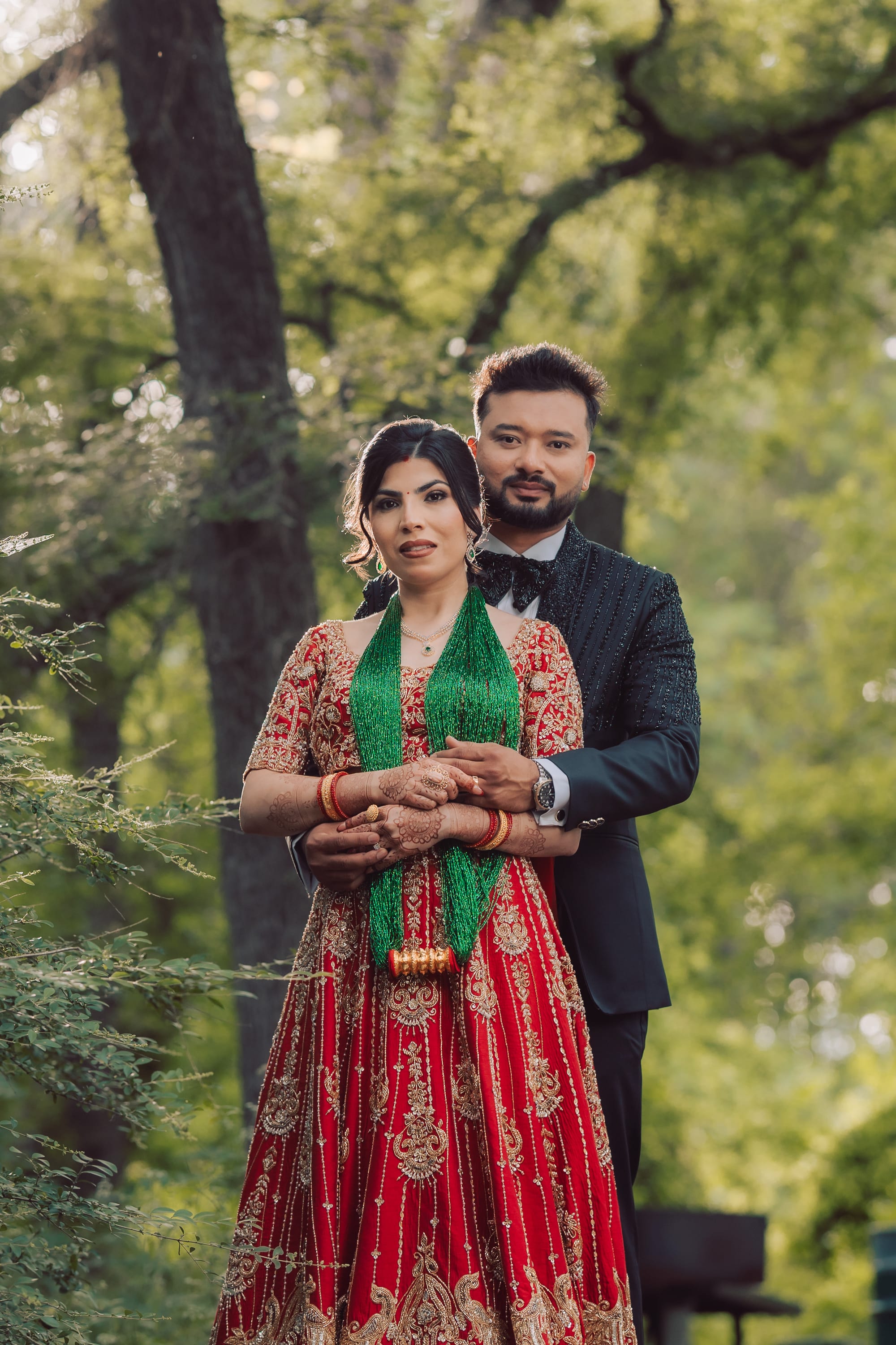 A groom in a beaded black tuxedo embraces a bride in a red and gold lehenga with a green beaded necklace among trees.