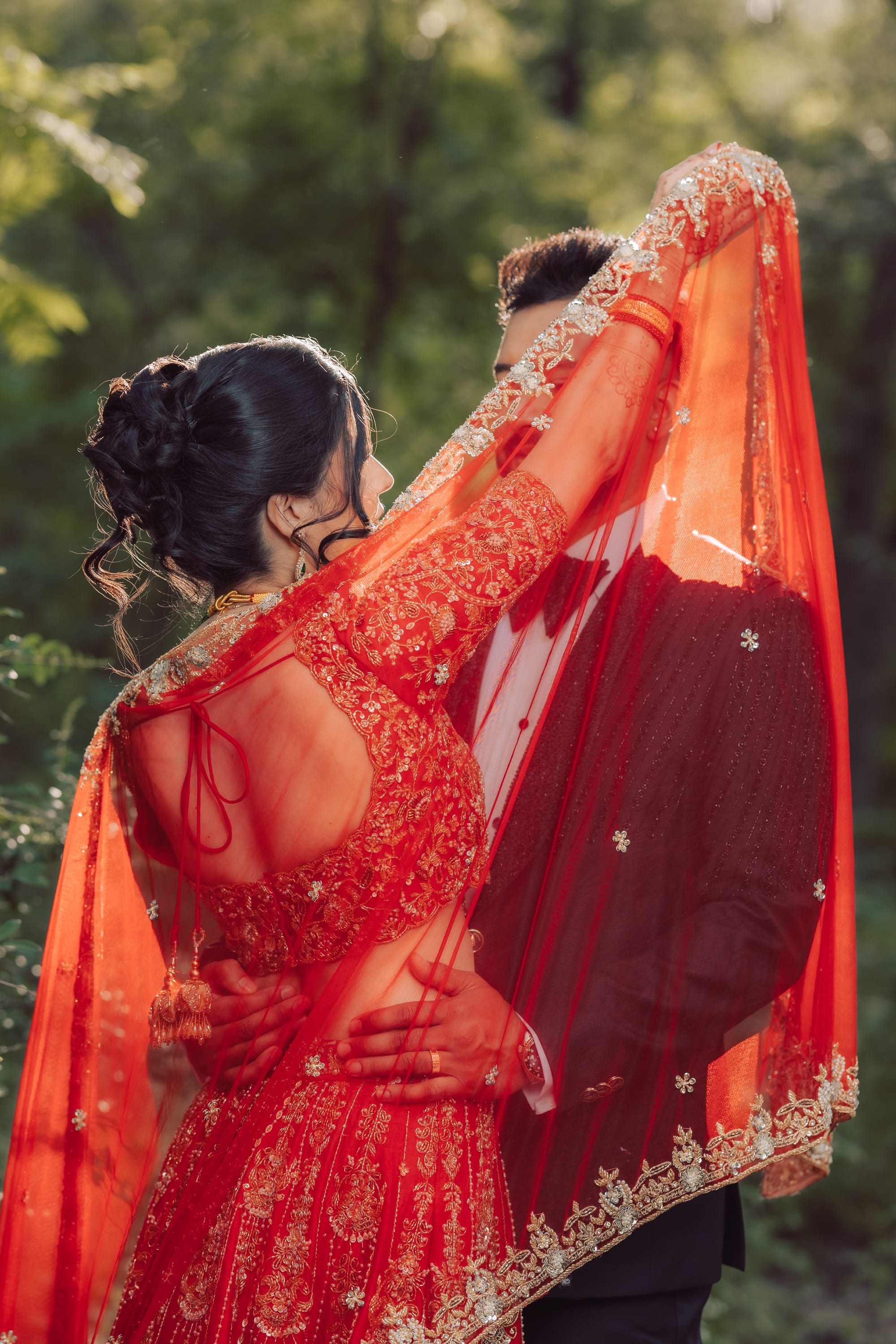 From behind, a bride in a red lehenga raises a sheer embroidered veil over herself and the groom in a forest.