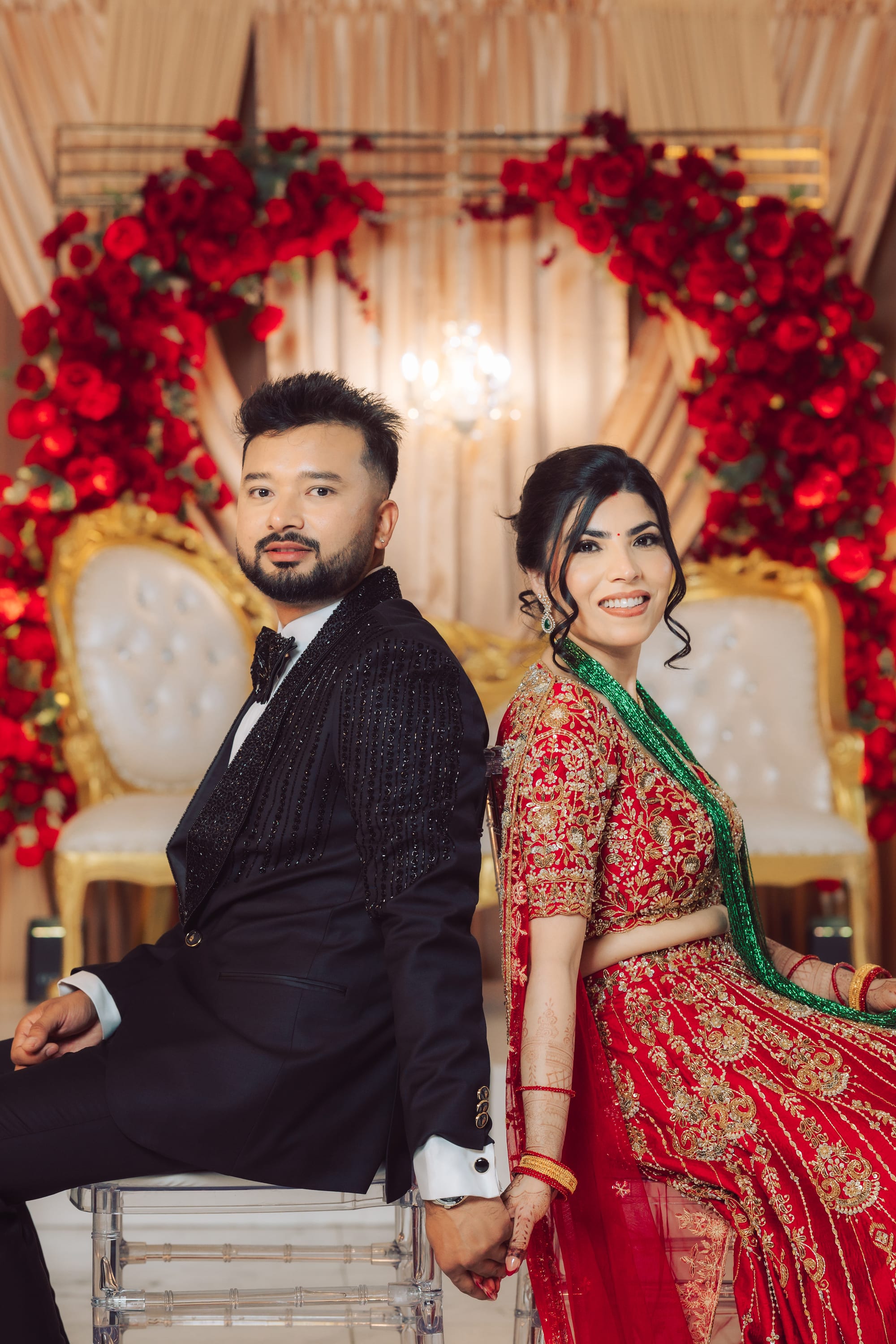 A bride in red and gold and a groom in a black tuxedo sit back to back, smiling, in front of a rose-covered arch.