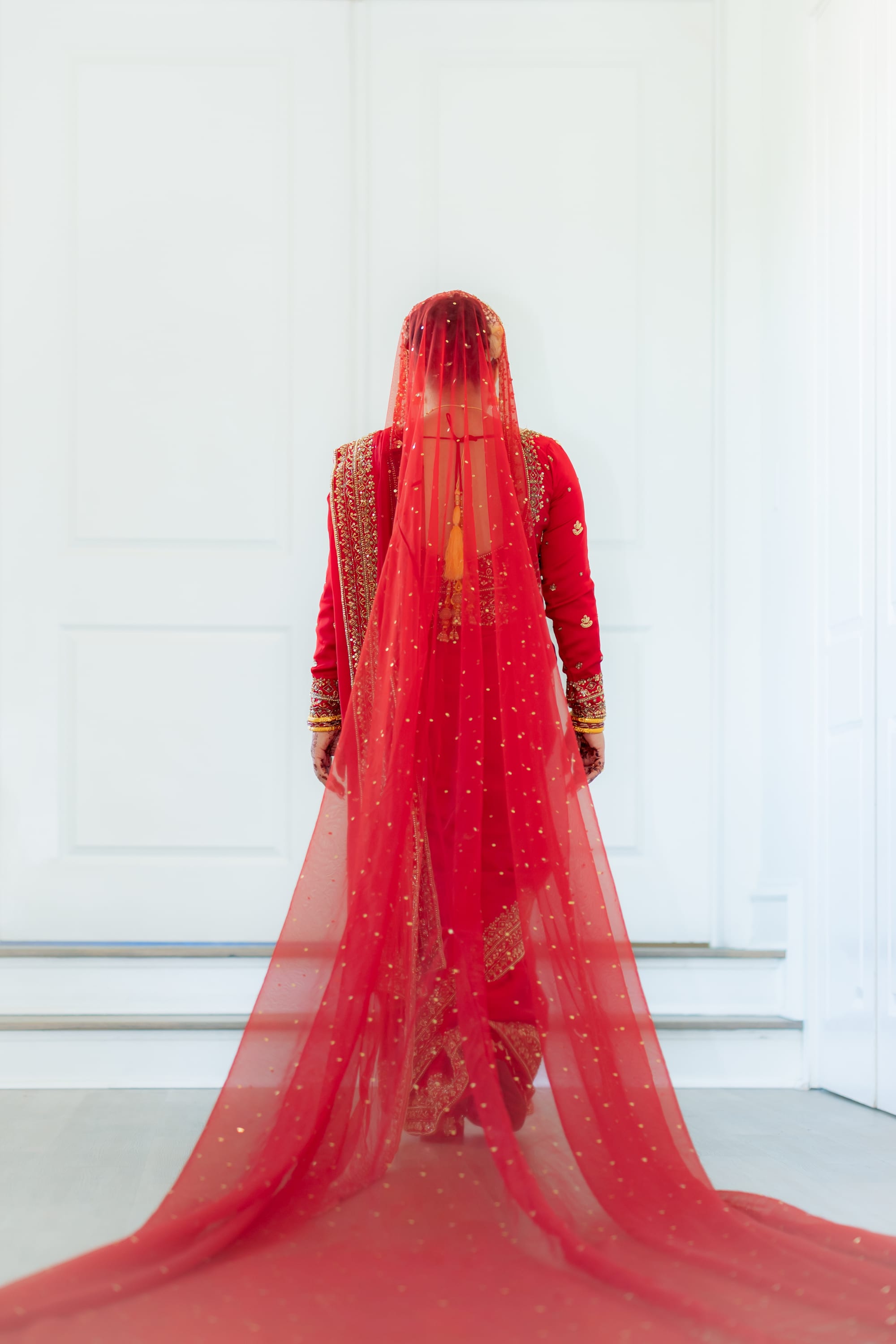 A bride seen from behind, wearing a long beaded red veil that trails across the floor.