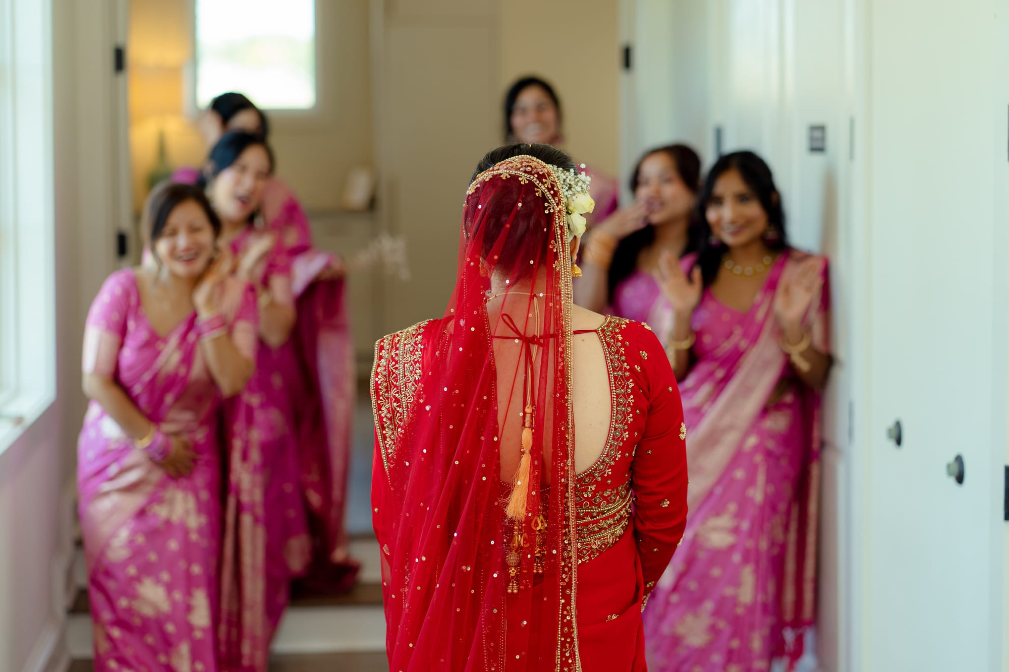 A bride in a red and gold veil walks toward bridesmaids in pink saris applauding her.