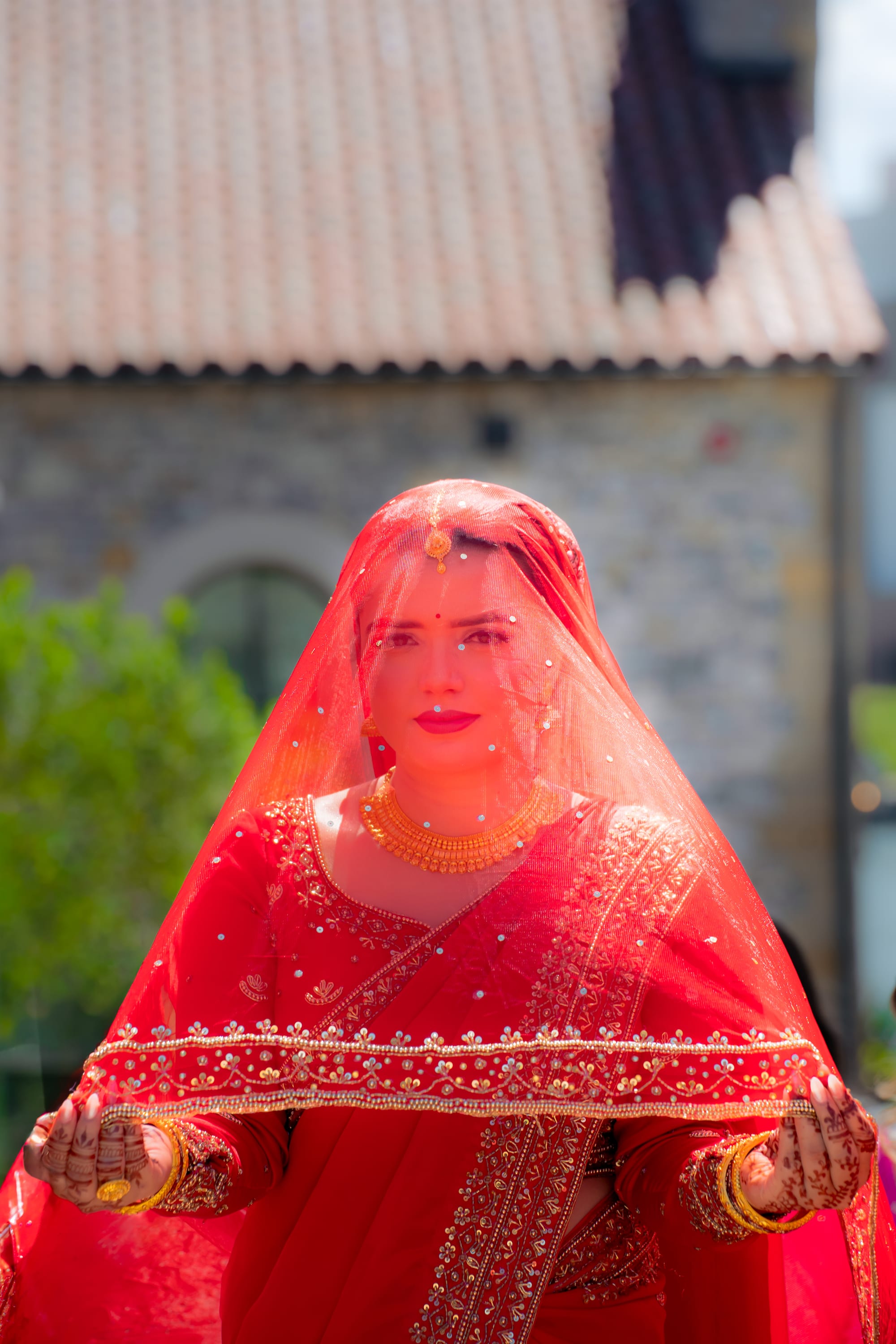 A bride in a sheer red veil and gold jewelry holds up the beaded edge of her dupatta.