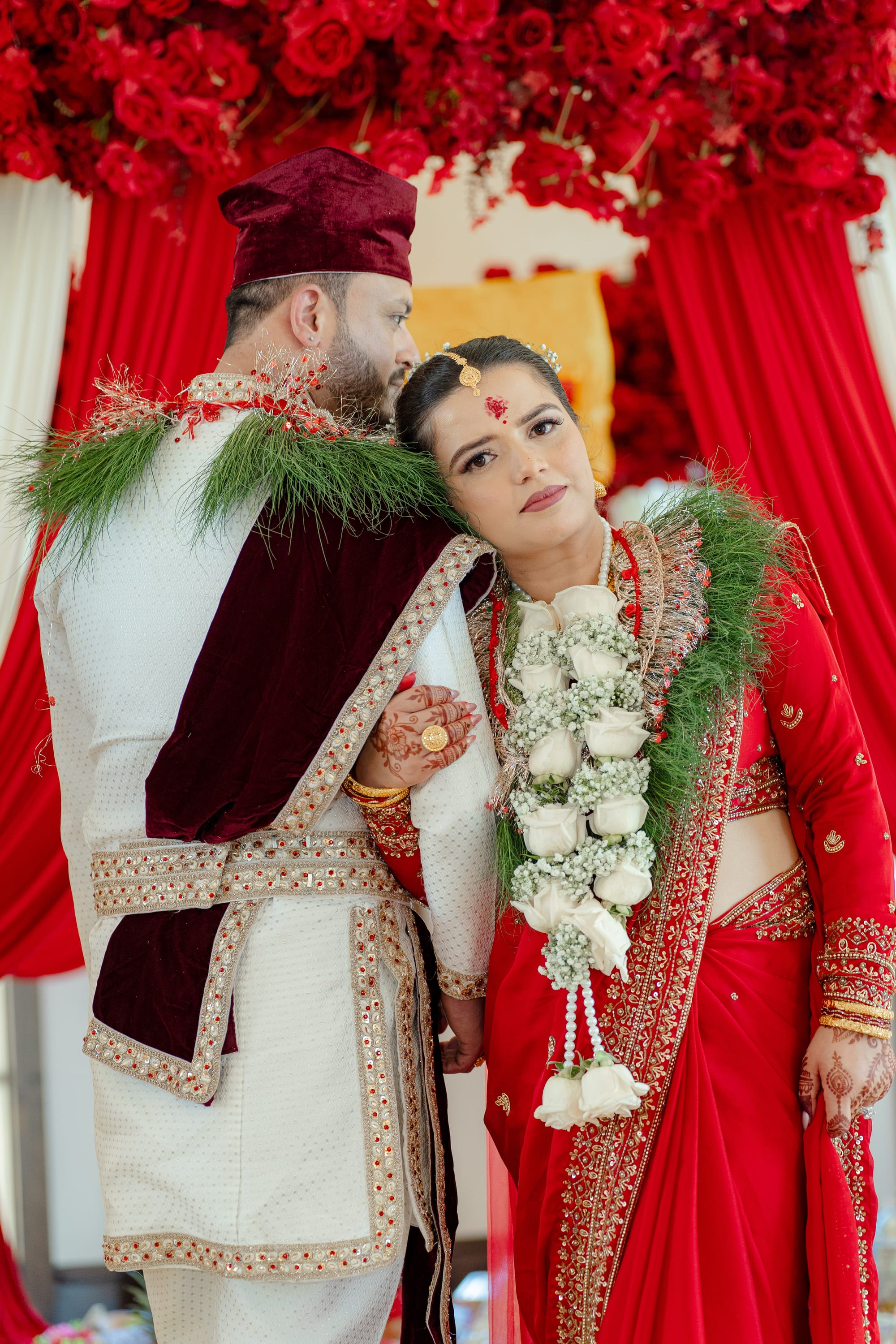 A bride rests her head on the shoulder of a groom in white and maroon, both wearing floral garlands.