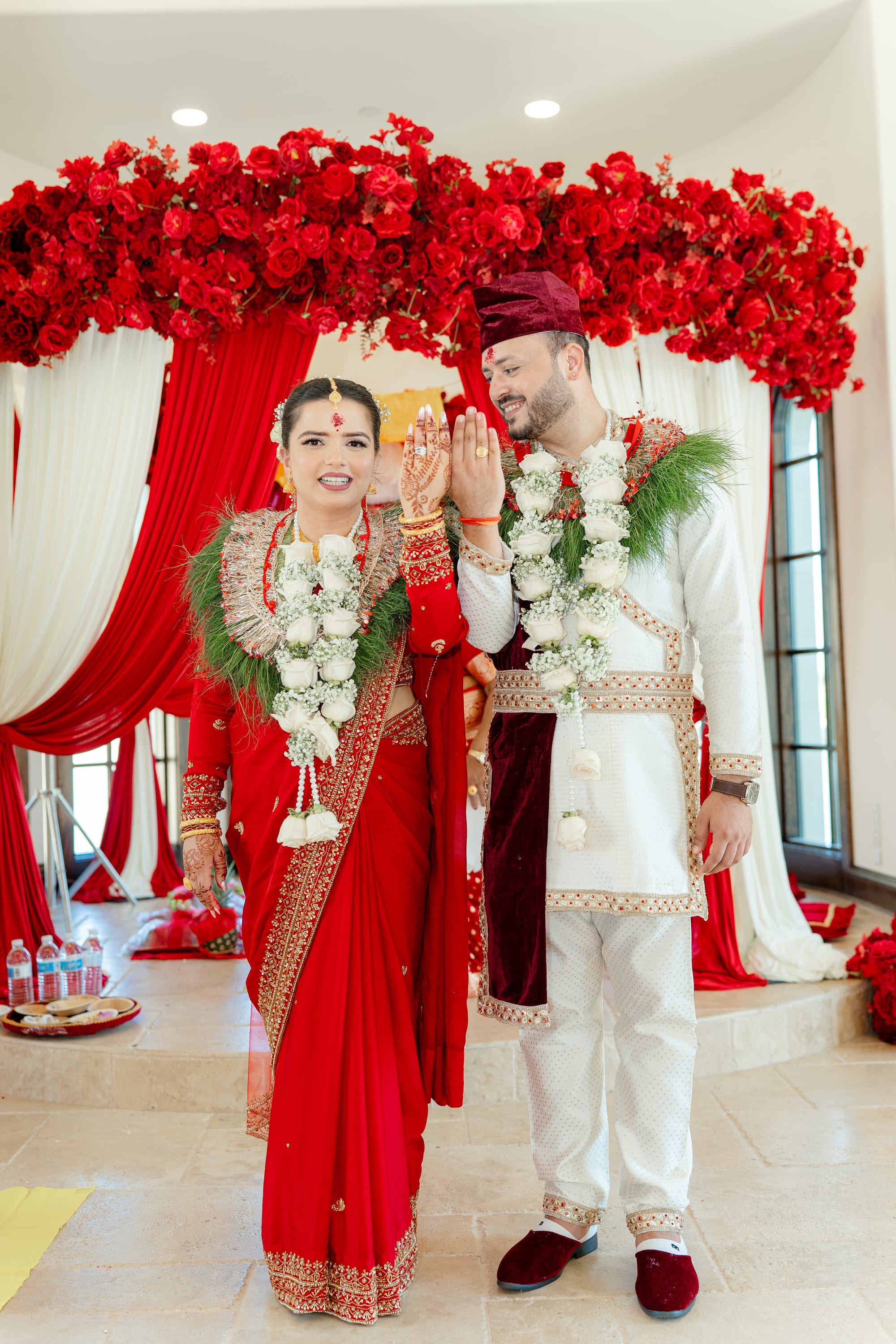 A bride and groom in red and white wedding attire with floral garlands raise their palms together, both smiling.