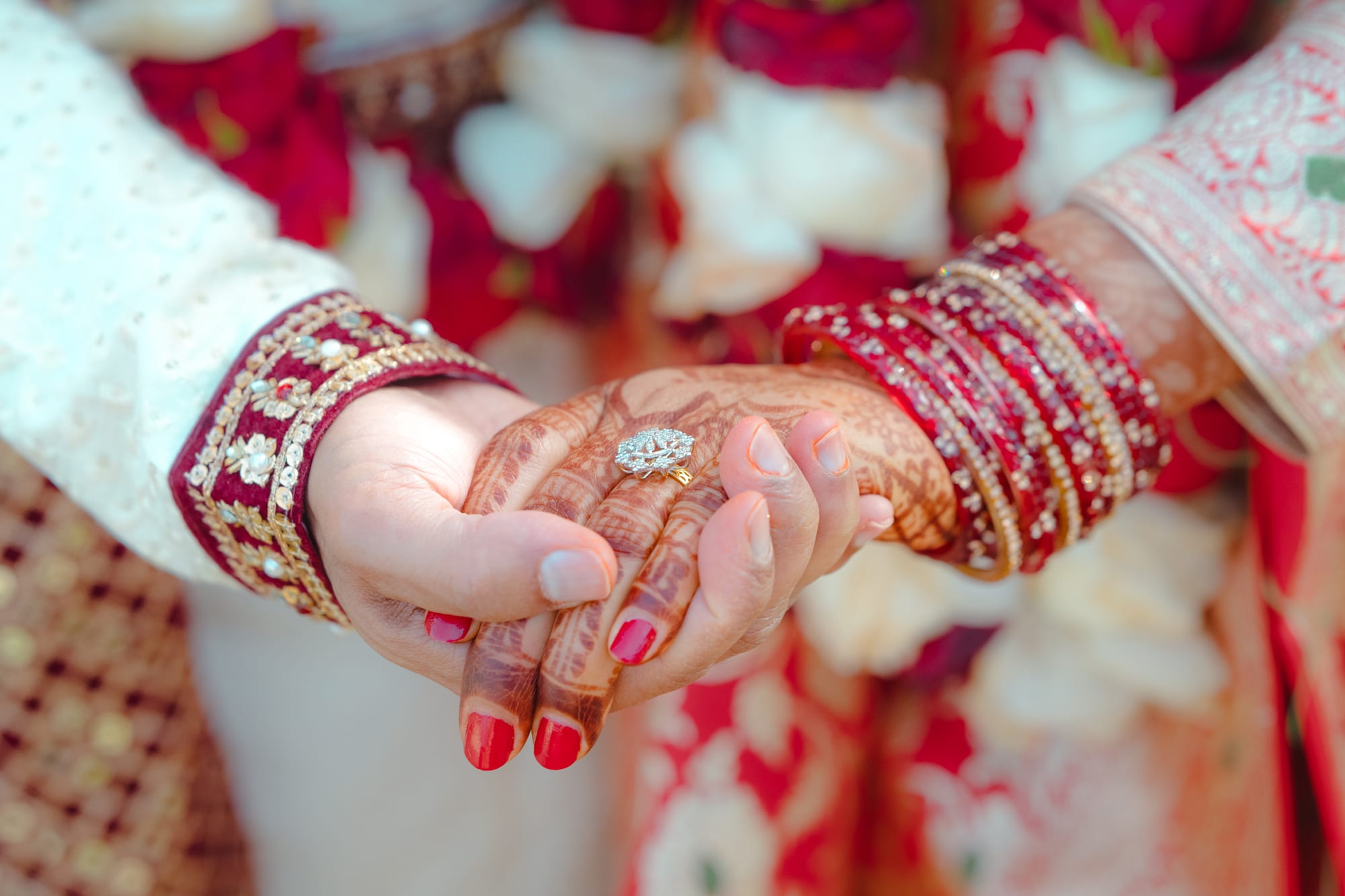 Close-up of a bride and groom's hennaed hands clasped together, framed by white floral garlands.