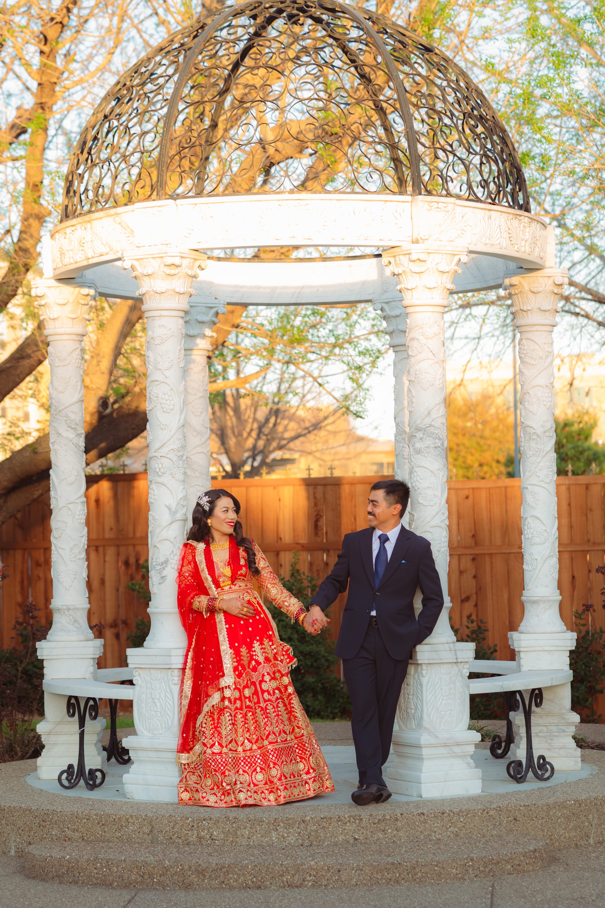 A bride in a red lehenga and a groom in a dark suit hold hands and smile at each other under an ornate white gazebo.