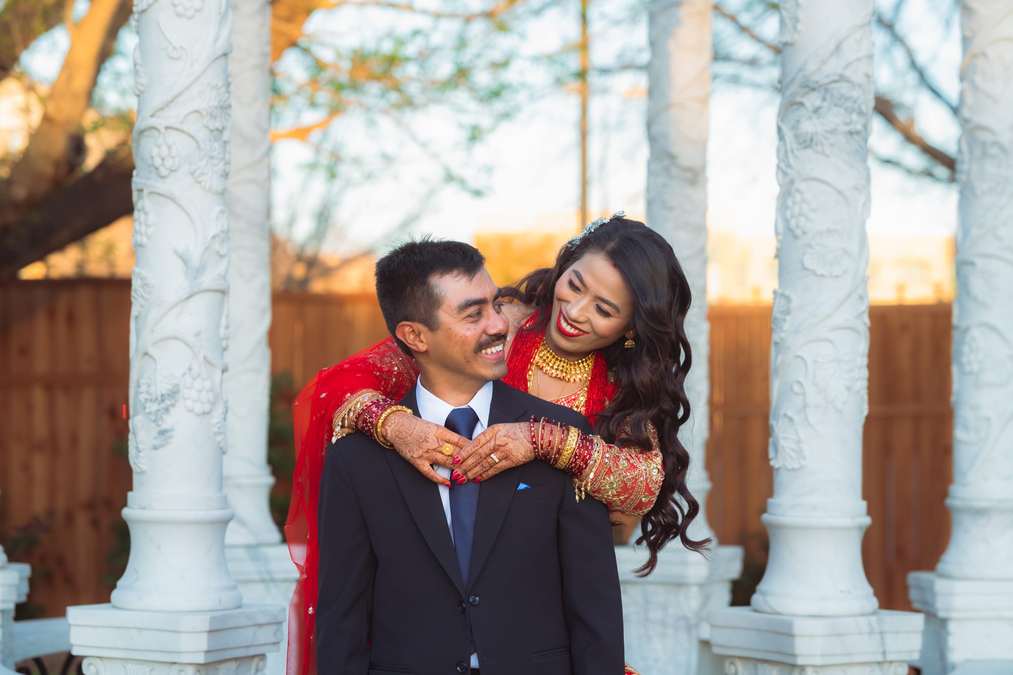 A bride in red with a sheer veil embraces a groom in a dark suit from behind, both smiling.